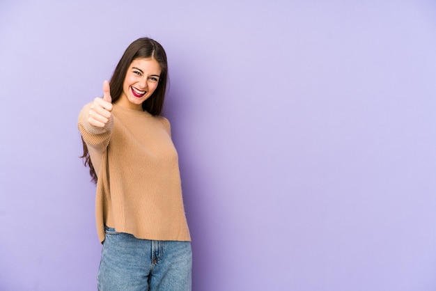 Mujer caucásica joven aislada en púrpura sonriendo y levantando el pulgar hacia arriba