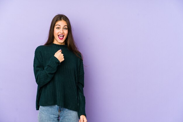 La mujer caucásica joven aislada en púrpura sorprendió señalar con el dedo, sonriendo ampliamente.