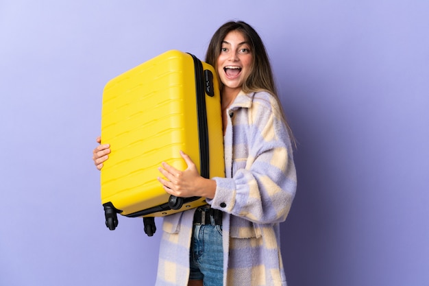 Mujer caucásica joven aislada en púrpura en vacaciones con maleta de viaje y sorprendida