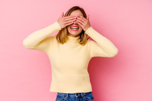 La mujer caucásica joven aislada en rosa cubre los ojos con las manos, sonríe ampliamente esperando una sorpresa.