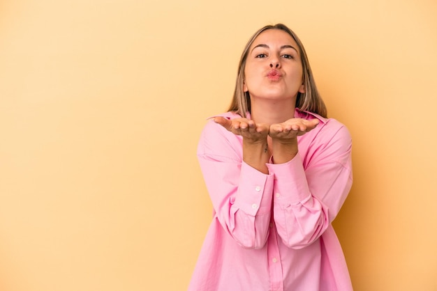 Mujer caucásica joven aislada sobre fondo amarillo doblando los labios y sosteniendo las palmas para enviar un beso al aire.