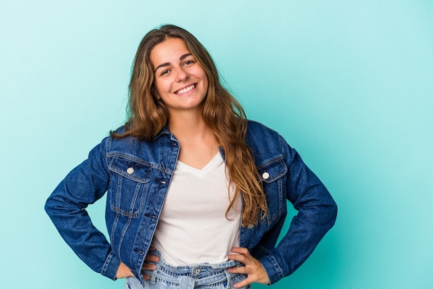 Mujer caucásica joven aislada sobre fondo azul confiado manteniendo las manos en las caderas.