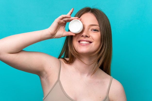 Mujer caucásica joven aislada sobre fondo azul con crema hidratante y felicidad