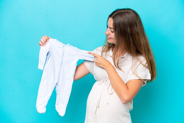 Mujer caucásica joven aislada sobre fondo azul embarazada y sosteniendo ropa de bebé