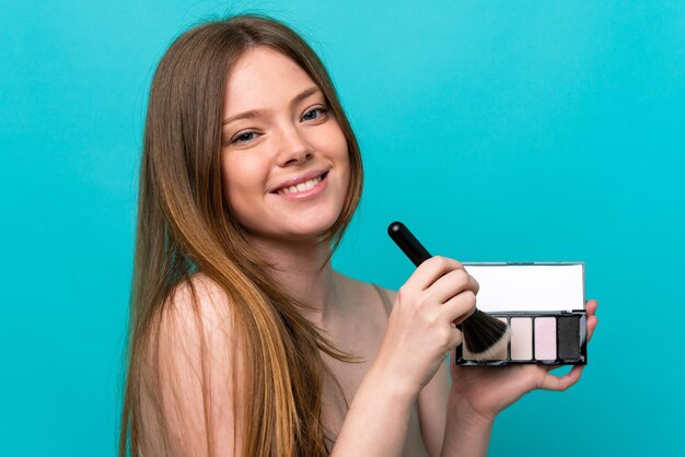 Mujer caucásica joven aislada sobre fondo azul con paleta de maquillaje y feliz