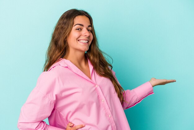 Mujer caucásica joven aislada sobre fondo azul que muestra una expresión de bienvenida.