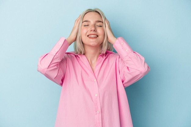 Mujer caucásica joven aislada sobre fondo azul se ríe alegremente manteniendo las manos en la cabeza Concepto de felicidad