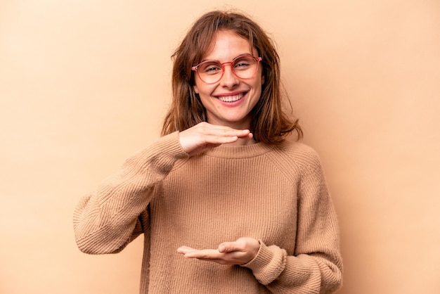 Mujer caucásica joven aislada sobre fondo beige sosteniendo algo con ambas manos presentación del producto