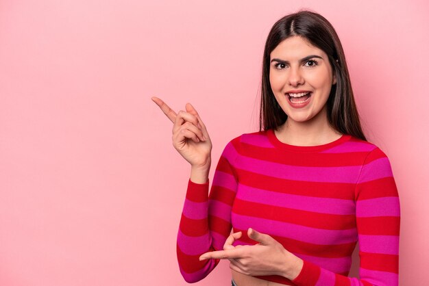 Mujer caucásica joven aislada sobre fondo rosa emocionada señalando con los dedos índices lejos