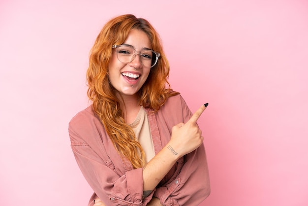 Foto mujer caucásica joven aislada sobre fondo rosa con gafas y lado señalador