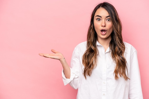 Mujer caucásica joven aislada sobre fondo rosa impresionada sosteniendo espacio de copia en la palma