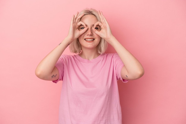 Mujer caucásica joven aislada sobre fondo rosa que muestra un signo bien sobre los ojos