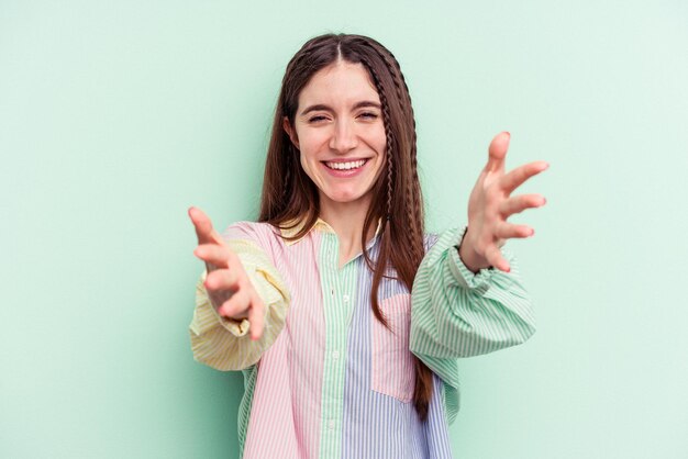 Foto mujer caucásica joven aislada sobre fondo verde que muestra una expresión de bienvenida.