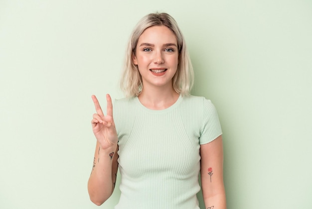 Mujer caucásica joven aislada sobre fondo verde que muestra el signo de la victoria y una amplia sonrisa.