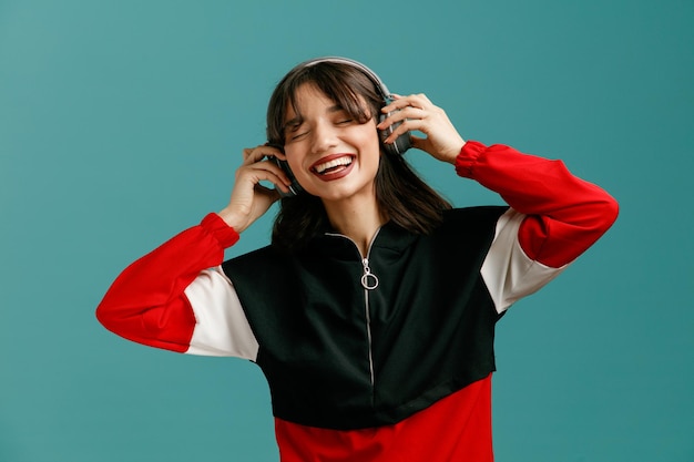 Mujer caucásica joven alegre usando auriculares agarrando auriculares disfrutando de la música con los ojos cerrados aislado sobre fondo azul.