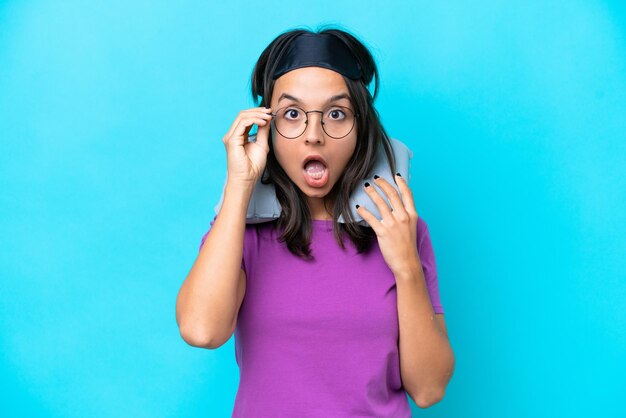 Mujer caucásica joven con almohada de viaje inflable aislada de fondo azul con gafas y sorprendida