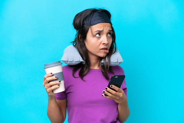 Mujer caucásica joven con almohada de viaje inflable aislada de fondo azul sosteniendo café para llevar y un móvil mientras piensa en algo