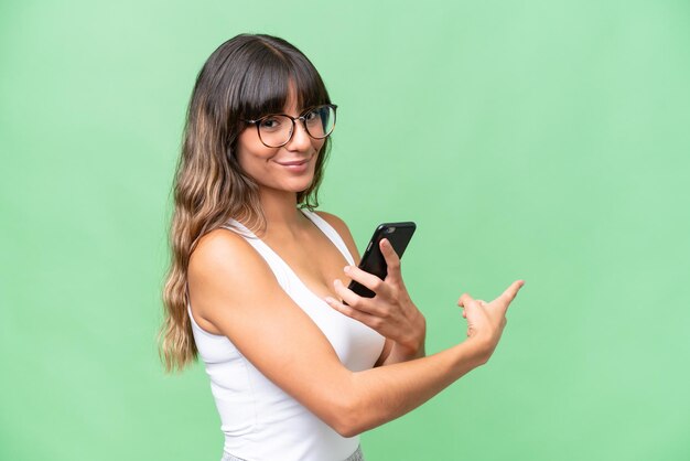 Mujer caucásica joven con antecedentes aislados usando un teléfono móvil y apuntando hacia atrás