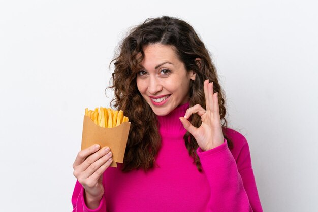 Mujer caucásica joven atrapando papas fritas aisladas en fondo blanco mostrando el signo de ok con los dedos