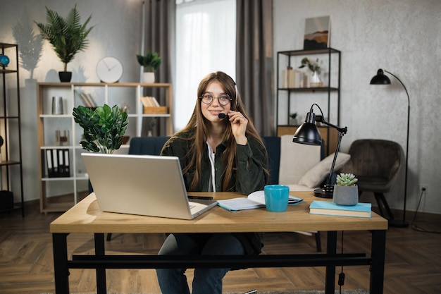 Mujer caucásica joven con auriculares sentada en el escritorio con una computadora portátil y hablando durante una videollamada