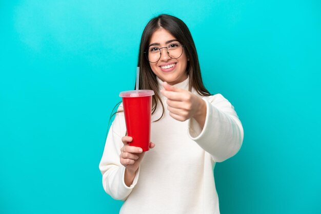 Mujer caucásica joven bebiendo refresco aislado sobre fondo azul haciendo gesto de dinero