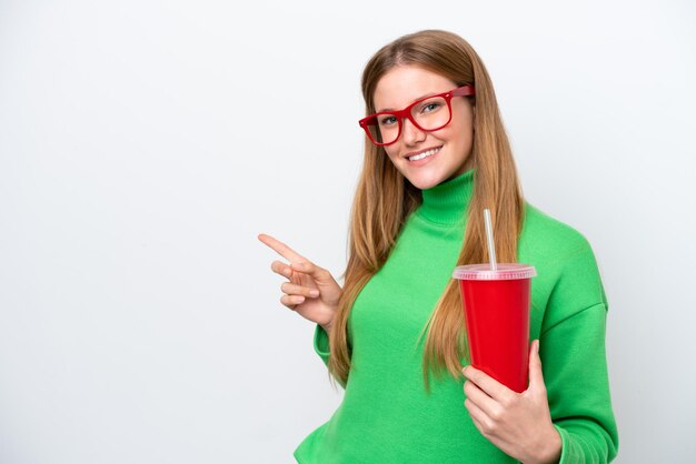 Mujer caucásica joven bebiendo refresco aislado sobre fondo blanco apuntando con el dedo al lado