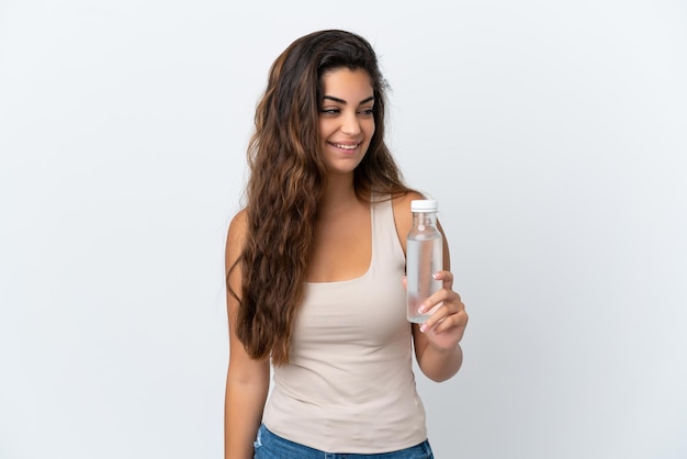 Mujer caucásica joven con una botella de agua aislada de fondo blanco mirando hacia un lado y sonriendo