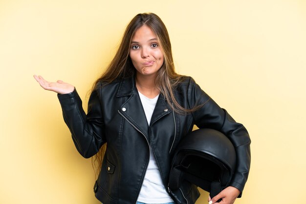 Mujer caucásica joven con un casco de motocicleta aislado en un fondo amarillo que tiene dudas mientras levanta las manos