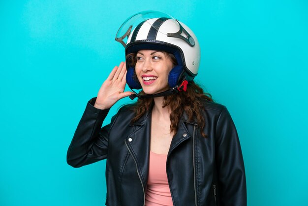 Mujer caucásica joven con un casco de motocicleta aislado de fondo azul escuchando algo poniendo la mano en la oreja