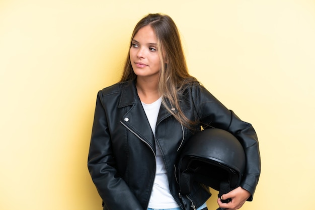 Mujer caucásica joven con un casco de motocicleta aislado sobre fondo amarillo mirando hacia el lado