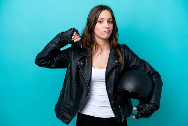 Mujer caucásica joven con un casco de motocicleta aislado sobre fondo azul que muestra el pulgar hacia abajo con expresión negativa