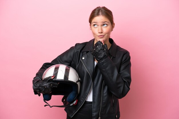 Mujer caucásica joven con un casco de motocicleta aislado sobre fondo rosa y mirando hacia arriba