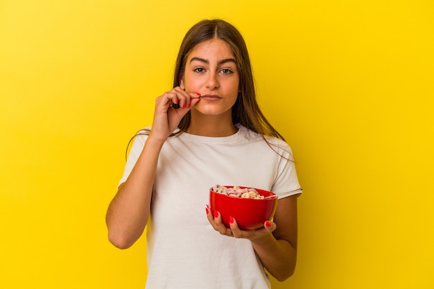 Mujer caucásica joven con cereales aislados sobre fondo amarillo con los dedos en los labios guardando un secreto.