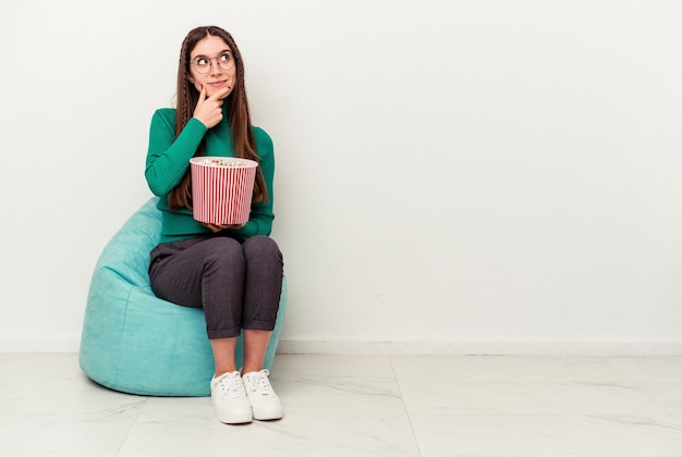 Mujer caucásica joven comiendo palomitas de maíz en un soplo aislado sobre fondo blanco mirando hacia los lados con expresión dudosa y escéptica