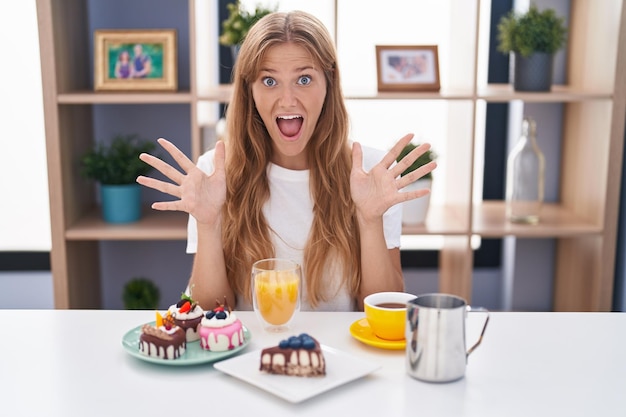 Mujer caucásica joven comiendo pasteles t para el desayuno celebrando loca y asombrada por el éxito con los brazos levantados y los ojos abiertos gritando emocionado concepto de ganador