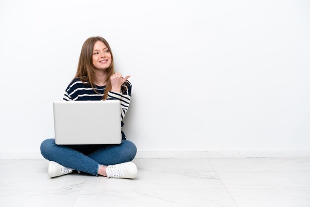 Mujer caucásica joven con una computadora portátil sentada en el suelo aislada de fondo blanco apuntando hacia un lado para presentar un producto