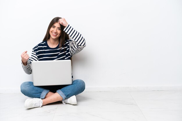 Mujer caucásica joven con una computadora portátil sentada en el suelo aislada de fondo blanco celebrando una victoria