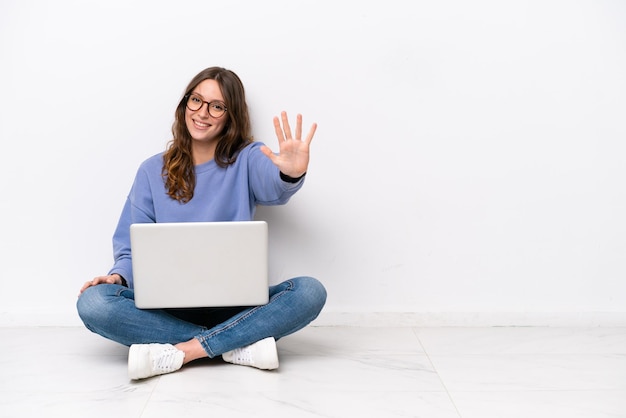 Mujer caucásica joven con una computadora portátil sentada en el suelo aislada de fondo blanco contando cinco con los dedos