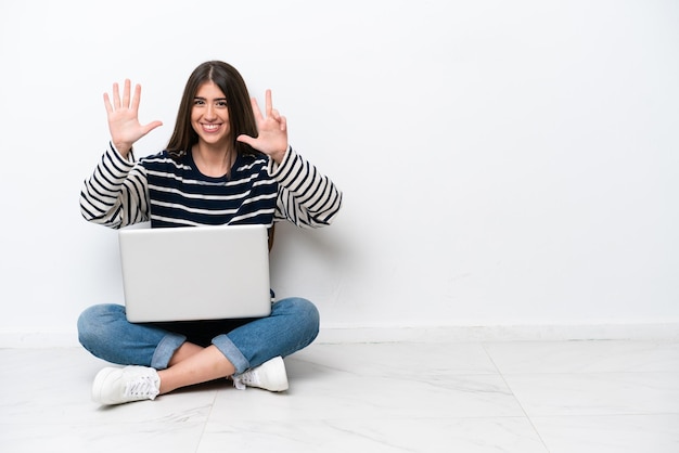 Mujer caucásica joven con una computadora portátil sentada en el suelo aislada de fondo blanco contando ocho con los dedos