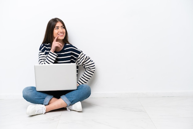 Mujer caucásica joven con una computadora portátil sentada en el suelo aislada de fondo blanco pensando en una idea mientras mira hacia arriba