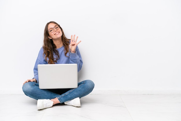 Mujer caucásica joven con una computadora portátil sentada en el suelo aislada de fondo blanco saludando con la mano con expresión feliz