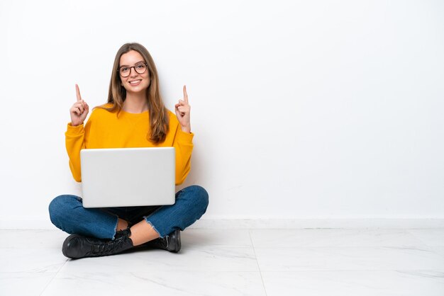 Mujer caucásica joven con una computadora portátil sentada en el suelo aislada de fondo blanco señalando una gran idea