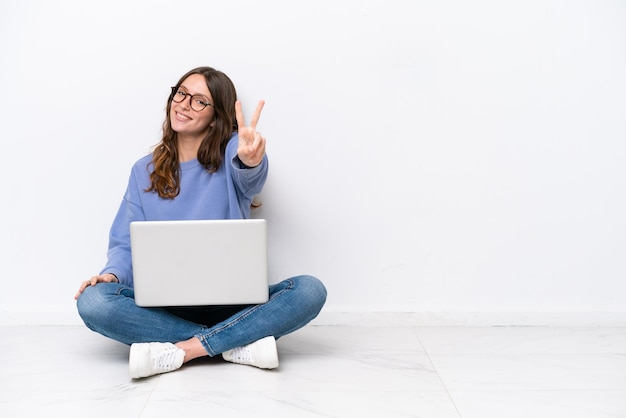 Mujer caucásica joven con una computadora portátil sentada en el suelo aislada de fondo blanco sonriendo y mostrando el signo de la victoria