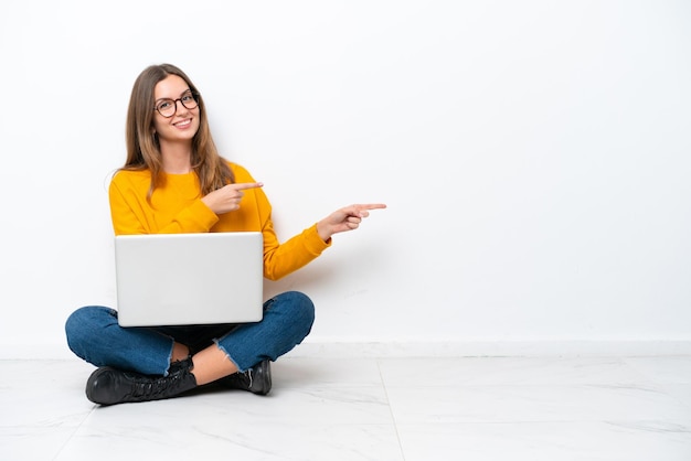 Mujer caucásica joven con una computadora portátil sentada en el suelo aislada de fondo blanco sorprendida y apuntando hacia el lado