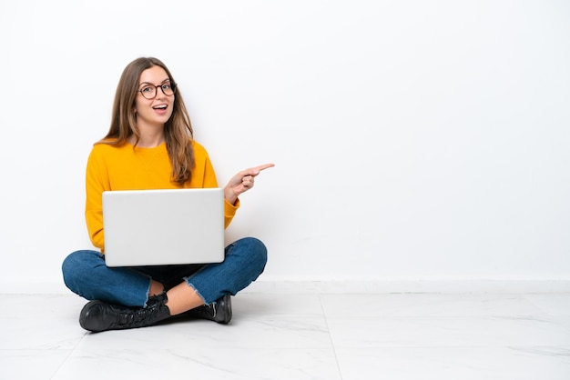 Mujer caucásica joven con una computadora portátil sentada en el suelo aislada de fondo blanco sorprendida y señalando con el dedo hacia un lado