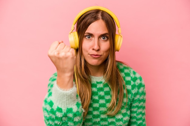 Mujer caucásica joven escuchando música aislada sobre fondo rosa mostrando el puño a la cámara expresión facial agresiva