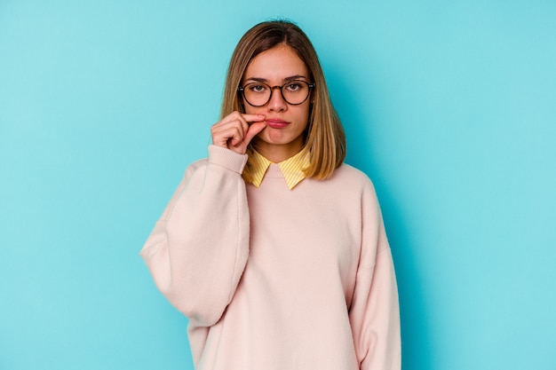 Mujer caucásica joven estudiante aislada en la pared azul con los dedos en los labios guardando un secreto.