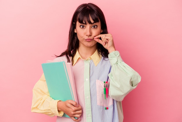 Mujer caucásica joven estudiante sosteniendo libros aislados sobre fondo rosa con los dedos en los labios guardando un secreto.