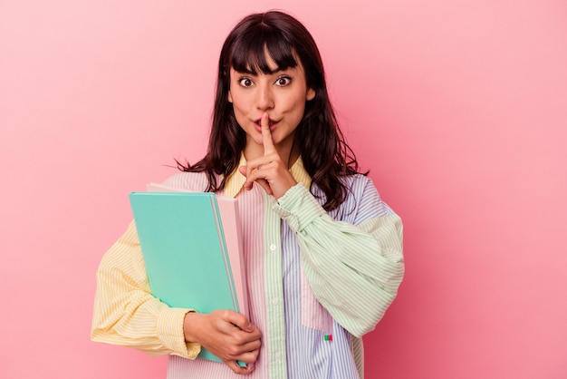 Mujer caucásica joven estudiante sosteniendo libros aislados sobre fondo rosa manteniendo un secreto o pidiendo silencio.