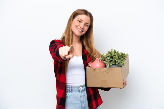 Mujer caucásica joven haciendo un movimiento mientras recoge una caja llena de cosas aisladas en fondo blanco apuntando al frente con expresión feliz